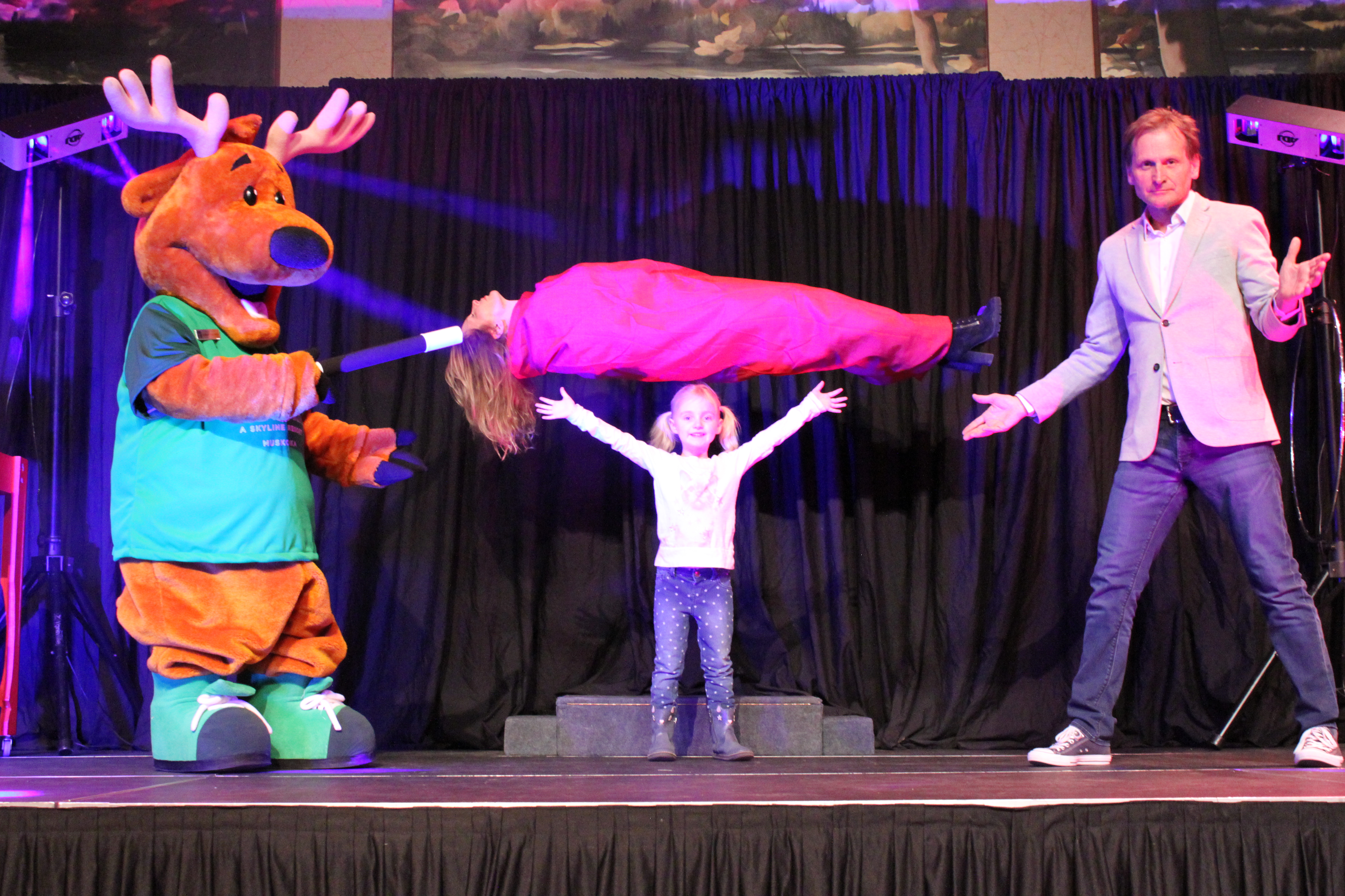 Magician Ron Gudel with young girl from audience and Charlie the Deer mascot on stage